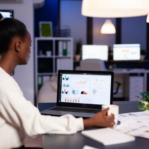 African businesswoman working with financial stats late at night in the office. Focused employee doing overtime for job respecting deadline of project writing, searching.