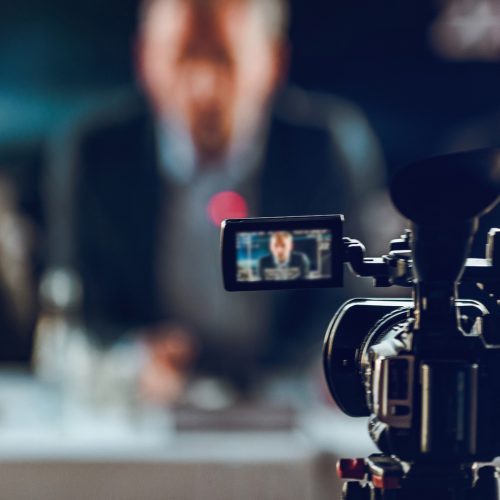 Professional digital camera at press conference, blurred speakers wearing suit background, live streaming concept