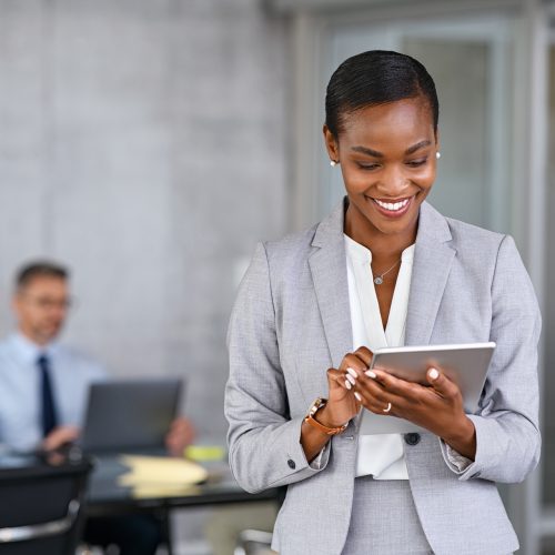Mature african businesswoman using digital tablet while standing in modern office. Black confident business woman working on digital tablet in modern office with copy sapce. Happy successful woman entrepreneur in formal clothing send email.