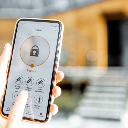 Holding phone with launched alarm app on the house background outdoors. Concept of controlling and managing home security from a mobile device