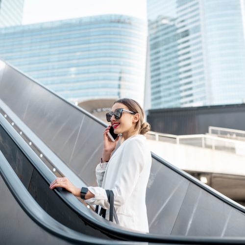 Business woman at the business center outdoors Stylish businesswoman in white suit talking phone while going up on the escalator at the business centre outdoors in Paris